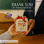 Person holding a 'Thank you Candle' box with a red ribbon, set against a blurred classroom background.